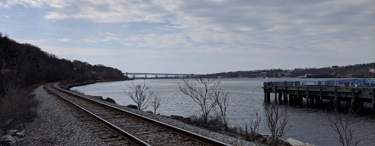 Train tracks run down the coastline towards a large bridge in the far distance. There is a dock on the right.